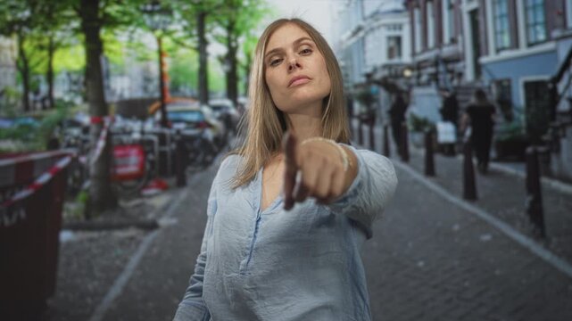 Young blonde woman stands amid parked bicycles on narrow cobblestone street flipping middle finger; defiance.