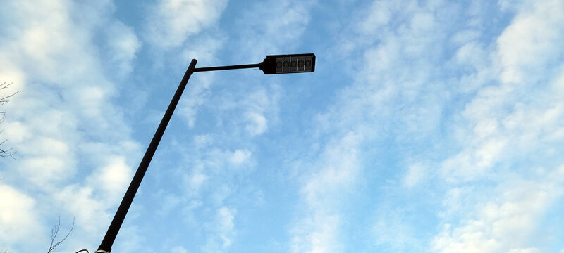 Modern LED street lamp stands tall against a bright, expansive blue sky filled with soft, white cirrocumulus clouds. The minimalist design of the black pole contrasts with the airy, natural background