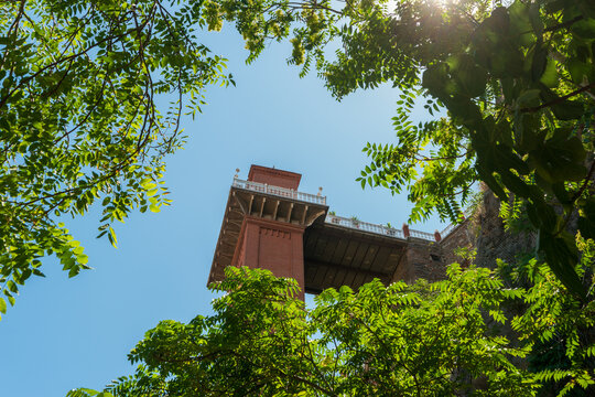 Izmir, Turkey. Historical Elevator Building tower's upper levels seen through green foliage with backlight, red brick contrasting with blue sky, low perspective from ground.