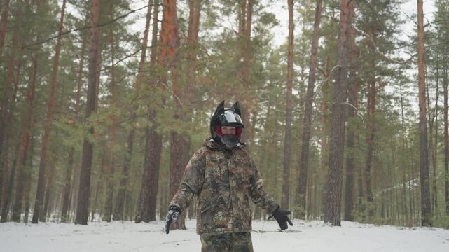 Helmeted rider stands in snowy forest, wearing camouflage jacket and red visor, light snow flurries fall among tall pine trunks, quiet winter atmosphere, steady stance as rider prepares for extreme