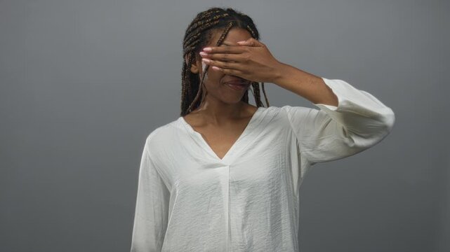 Woman with hand covering eyes, braided hair, long nails and white blouse in a gray studio; shyness introspection reflection.