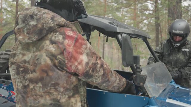 Camo riders prepare atv in forest, two people in camouflage jackets approach blue utv in snowy pine woods, check windshield and gear, strap helmets, test controls, passenger climbs into seat, quick