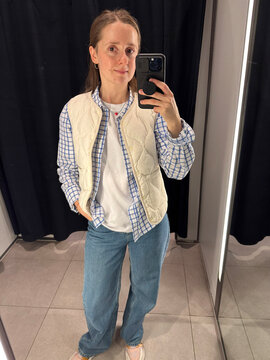 Young woman taking a mirror selfie while trying on clothes in a fitting room. Modern shopping experience, fashion choices and consumer lifestyle concept