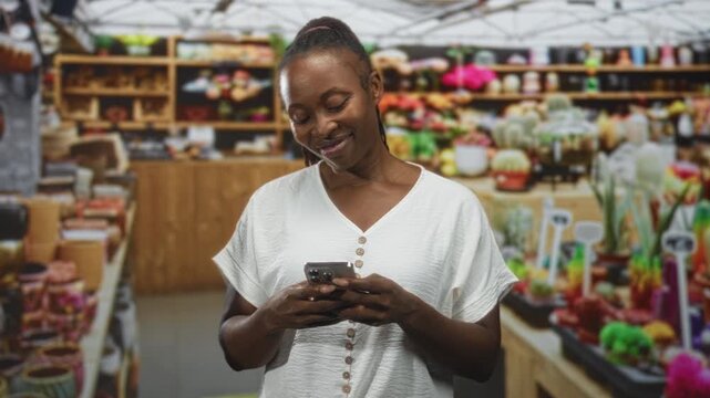 Young africanamerican woman holding a smartphone, smiling and texting inside a building market with shelves of pots, succulents and wooden displays; contentment connection.