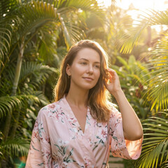 Calm young woman portrait among tropical nature enjoying bright sunlight. She wears pink floral robe with serene emotional expression surrounded by green palm leaf touching her beautiful hair