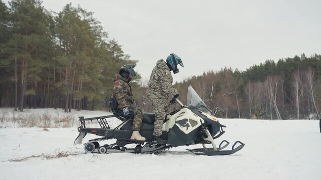 Two riders on snowmobile preparing gear in snowy clearing near pine forest, camouflage suits and helmets, one seated on vehicle while other adjusts equipment, breath visible in cold air, quiet offroad