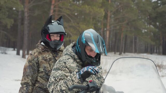 Two riders inspect atv before departure, camouflage coats and helmets with visors, snowy forest clearing, driver checks windshield and throttle, passenger reviews checklist, teamwork briefing, gear