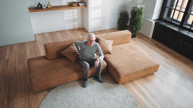 Sick senior man rubbing knee when sitting on couch in living room, ache in joint . Aging and disease of old age, stay healthy and active after 70 years, arthritis of knee joint or trauma, feel pain