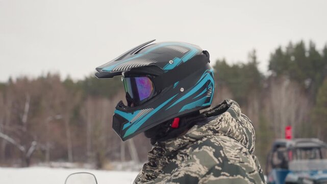 Closeup snowmobile helmet with mirrored goggles, teal and black graphics, visor reflection of winter trees, camo jacket collar, protective padding detail, product style focus on safety gear