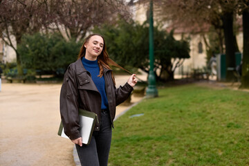 A woman in a brown leather jacket and blue turtleneck stands in a park, touching her hair while holding a laptop in a green sleeve. © dsbedarieva