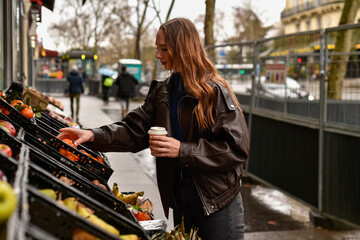 A woman in a brown leather jacket holds a coffee cup while selecting tangerines from an outdoor fruit display on a city street in Paris. © dsbedarieva