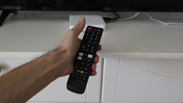 Male hand brings a TV remote control into frame, presses the power button, and places it on a cabinet in front of the television next to a media box. Concept of turning on TV and home entertainment se