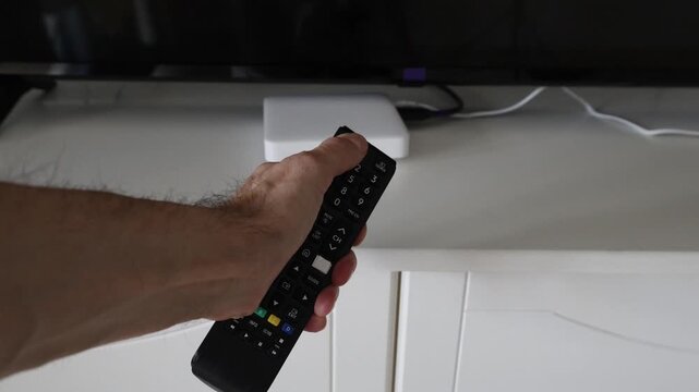 Male hand picks up a TV remote control from a cabinet, presses the red power button, and places the remote back. Media box and television visible in the background. Concept of turning TV on and home e