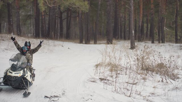 Snowmobile joy outdoors. Adventurous snowmobiling through dense pine forest during winter day. Thrillseeker in camouflage gear raising arms while carving through snowy wilderness landscape