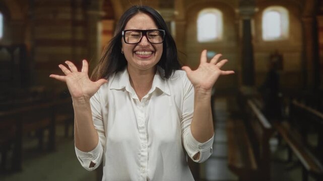 Woman with hands raised smiling and palms open inside building church pews, raises hands in praise; joy faith celebration.