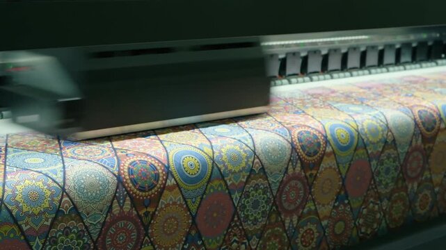 Large-format printer moves across colorful patterned fabric. Print head advances steadily, depositing vivid hues onto material. Pattern repeats geometrically