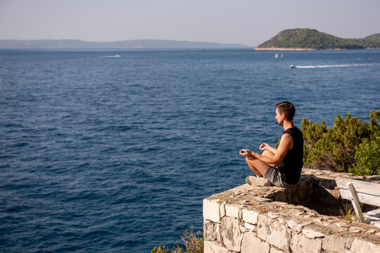 Man meditating in yoga pose on rocky ledge by the sea