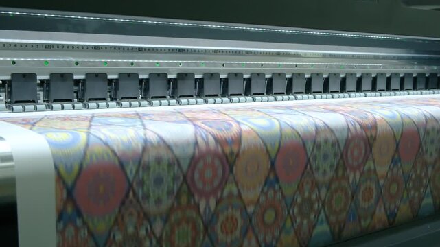 Large-format printer moves across colorful patterned fabric. Print head advances steadily, depositing vivid hues onto material. Pattern repeats geometrically