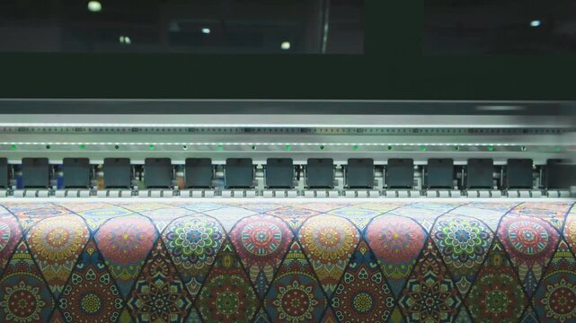 Large-format printer moves across colorful patterned fabric. Print head advances steadily, depositing vivid hues onto material. Pattern repeats geometrically