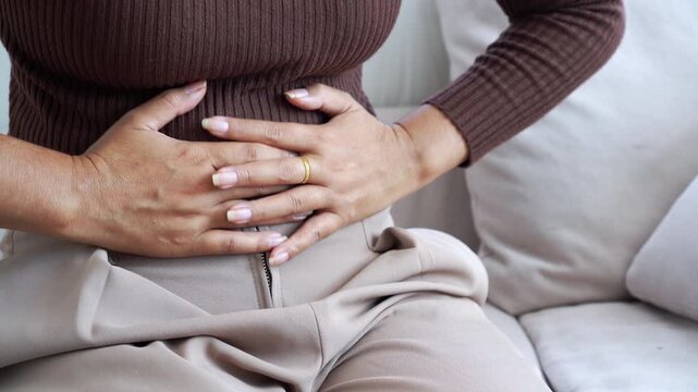 Close-up of a woman clutching her abdomen with both hands, suffering from severe stomach ache, food poisoning, or menstrual cramps at home. Concept of medical health problems and indigestion