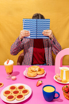 Person hiding face with book at colorful breakfast table