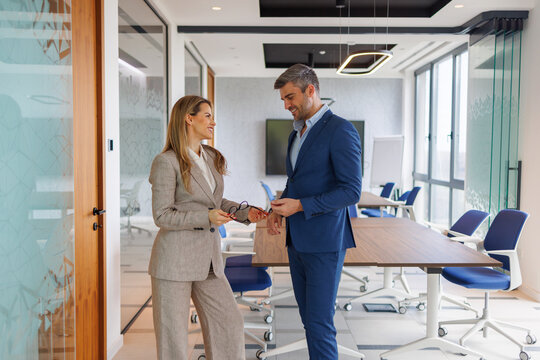 Two business professionals in business attire stand and conversing in a bright, contemporary office meeting room
