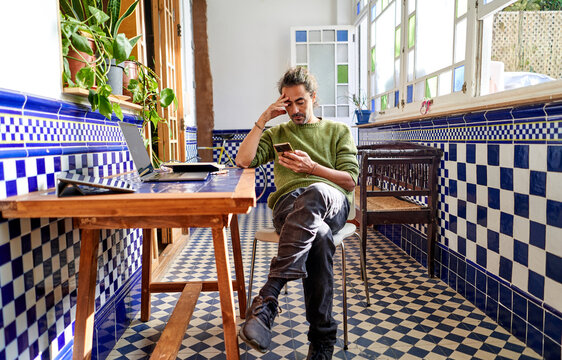 Hipster middle aged man using phone and laptop at home table