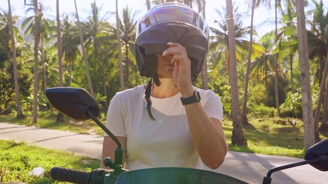 Young Woman Sitting on Scooter Lifting Front Part of Protective Helmet While Standing on Tropical Road With Palm Trees and Bright Sunlight, Travel Pause and Comfort Adjustment