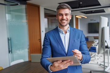 Fototapeta premium Confident businessman smiling, holding a tablet while standing in a modern office, representing success and technology