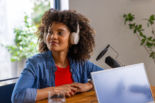 African American woman sitting in home studio wearing shirt and headset recording on mic and laptop