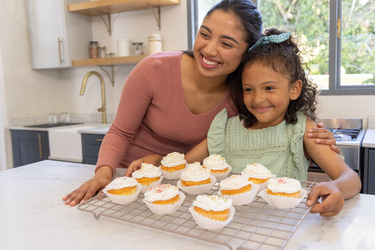 Mother and child smiling and presenting frosted cupcakes on cooling rack on marble countertop