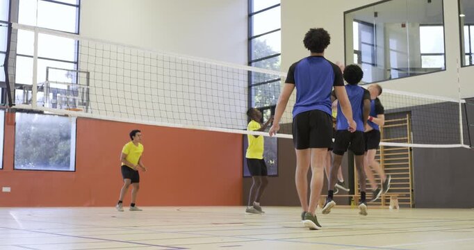 Six male volleyball players rallying at school gym after serve, blue bib player attacking net