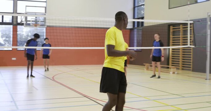 Yellow-shirted male team retrieving multicolored ball in school gym, serving to continue rally