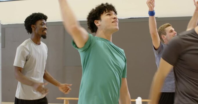 Diverse male teammates heading to volleyball net in gym after scoring point, high-fiving near bench