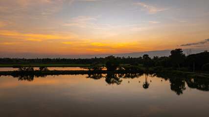 Obraz premium Scenic sunset sky glowing with orange and golden tones above a tranquil wetland. Reflections on the still water and dark tree silhouettes create a serene countryside atmosphere in this beautiful