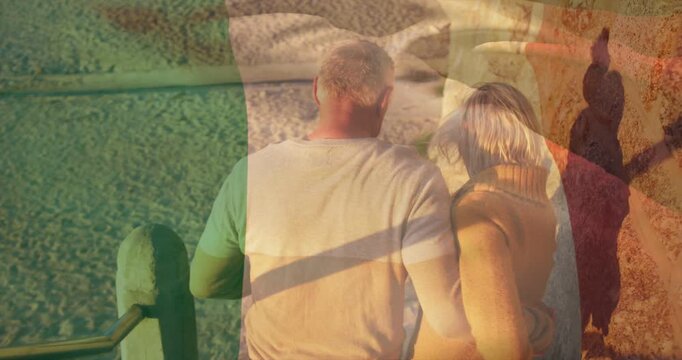Walking older couple down coast stairs to beach holding metal railing, wearing tan sweater, overlay