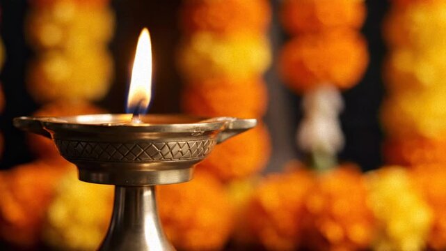 A glowing kuthuvilakku oil lamp in a warm close-up, with blurred festive marigold garlands for puthandu.