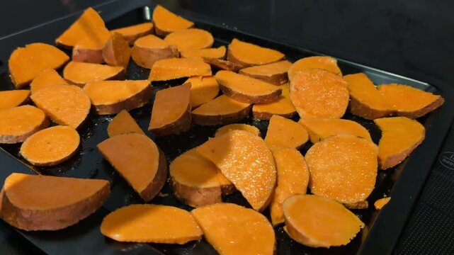 Sliced sweet potatoes roasting on baking tray in oven, Close-up of seasoned orange root vegetable pieces cooking on metal sheet pan with oil, prepared for a homemade healthy dish
