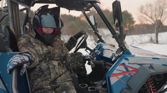 Snowy utv rider readies atv controls wearing camo jacket fullface helmet and gloves sits inside blue utility vehicle hands on steering and throttle dusk light snow covered field distant trees tense