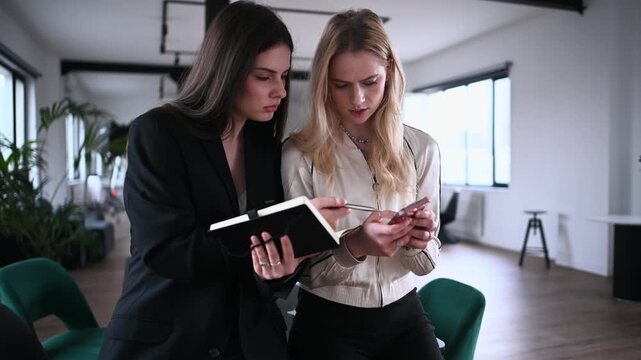 Young businesswomen collaborating on a project in a modern office, one showing information on her smartphone while the other takes notes in a planner, highlighting teamwork and communication