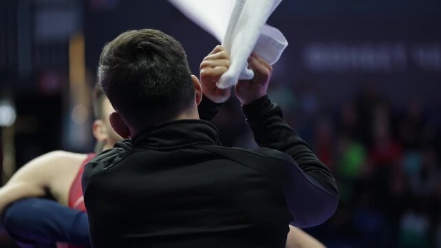 Back-view official oversees an intense indoor wrestling bout as athletes grapple on the mat, with a blurred crowd and arena lighting framing a dramatic sideline perspective.