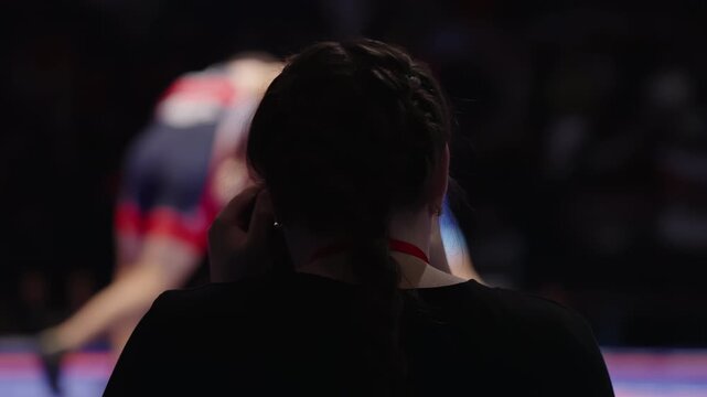 Silhouetted spectator with braided hair watches an intense wrestling bout under arena lights, focusing on the grappling athletes as they struggle on a colorful mat in the blurred background