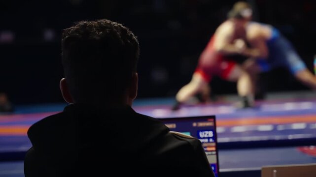 Silhouetted official monitoring a wrestling bout from a table as two athletes grapple on an illuminated mat, with scoreboard and laptop screens glowing in a dimly lit indoor arena.