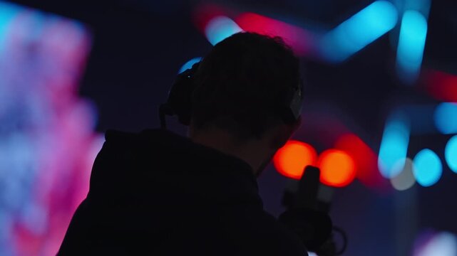 Silhouette of a camera operator filming on a vibrant stage with colorful bokeh lights and neon shapes, capturing live event action in a dark venue with dramatic backlighting.