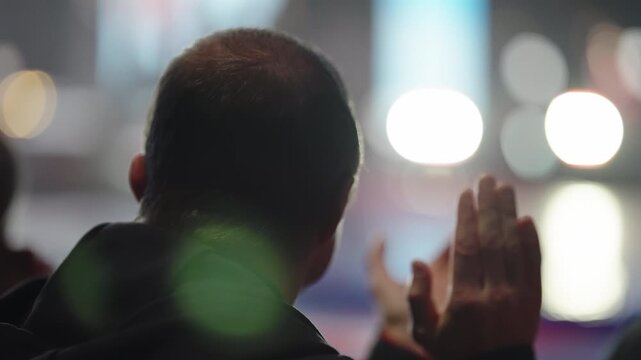 Audience member applauding during a live performance, viewed from behind with soft bokeh stage lights and low-light mood, capturing the closing moments of an engaging event.
