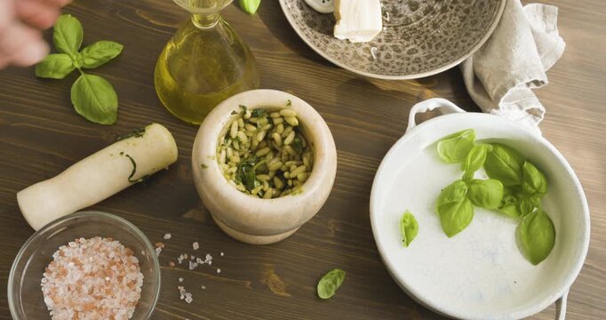 Person places glass carafe of extra virgin olive oil on the table. Marble mortar for Genoese pesto. Aerial view: preparing traditional Italian sauce on rustic wooden table under natural sunlight