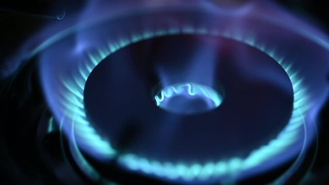 Close up macro shot of a bright blue gas flame igniting and burning on a kitchen stove burner ring against a dark background