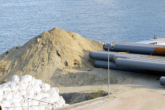 Riverside construction site with a large sand pile, gray pipes, and bulk bags against the blue water. Shows civil engineering material staging, preparation activity, and large-scale project supply.