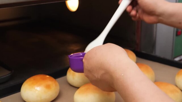 A close-up shot of a person using a silicone brush to apply a shiny egg wash or butter glaze to warm, golden-brown dinner rolls on a baking sheet. The scene captures the final steps of baking soft, ho