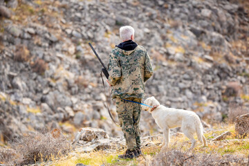 Naklejka premium A hunter in camouflage with a rifle stalks game in the mountains. Hunting for game in the mountains. A man with a rifle and dog practices hunting and shooting together in the wild.